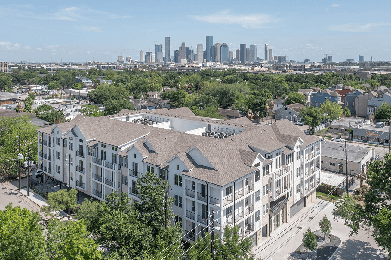 Aerial view of a four-story apartment community set among tree-lined streets with the downtown skyline beyond
